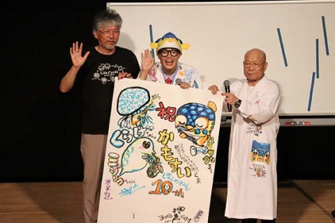 さかなクン（中央）が描いたリニューアル10周年の絵を持って坂田さん（右）と奥泉館長と共に会場に集まった人たちの記念撮影に応じた