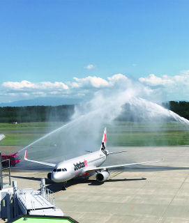 夏空が広がる庄内空港にジェットスターの初便が降り立ち、駐機場では消防車の放水アーチで就航を歓迎