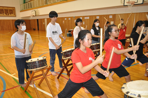 辨天太鼓メンバー（前列）のたたき方を参考に、太鼓演奏に挑戦する曽於市の小中高生＝2日