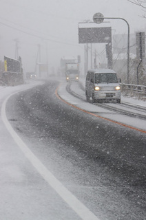 雪が降りしきる中、ヘッドライトをつけて走る車＝25日午前10時過ぎ、鶴岡市田麦俣の国道112号