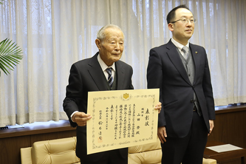 長年にわたる町内会長としての功績で総務大臣表彰を伝達された山田さん（左）＝鶴岡市役所