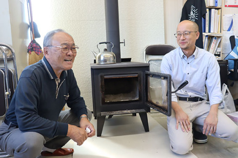 「今年も出番が近づいてきたな」―。薪ストーブの前で談笑する加藤自治会長（左）と竹内事務局長＝三瀬コミセン