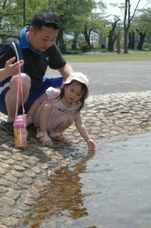 強い日差しの下、疎林広場の水場で水とたわむれる親子